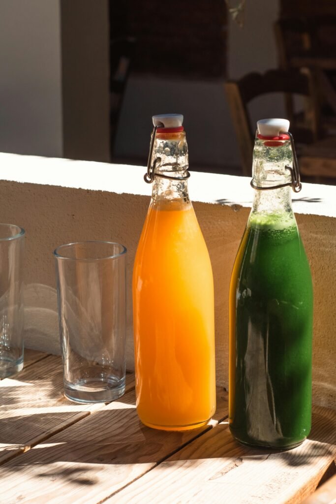 Two glass bottles filled with orange and green juices sitting on a sunlit wooden table.