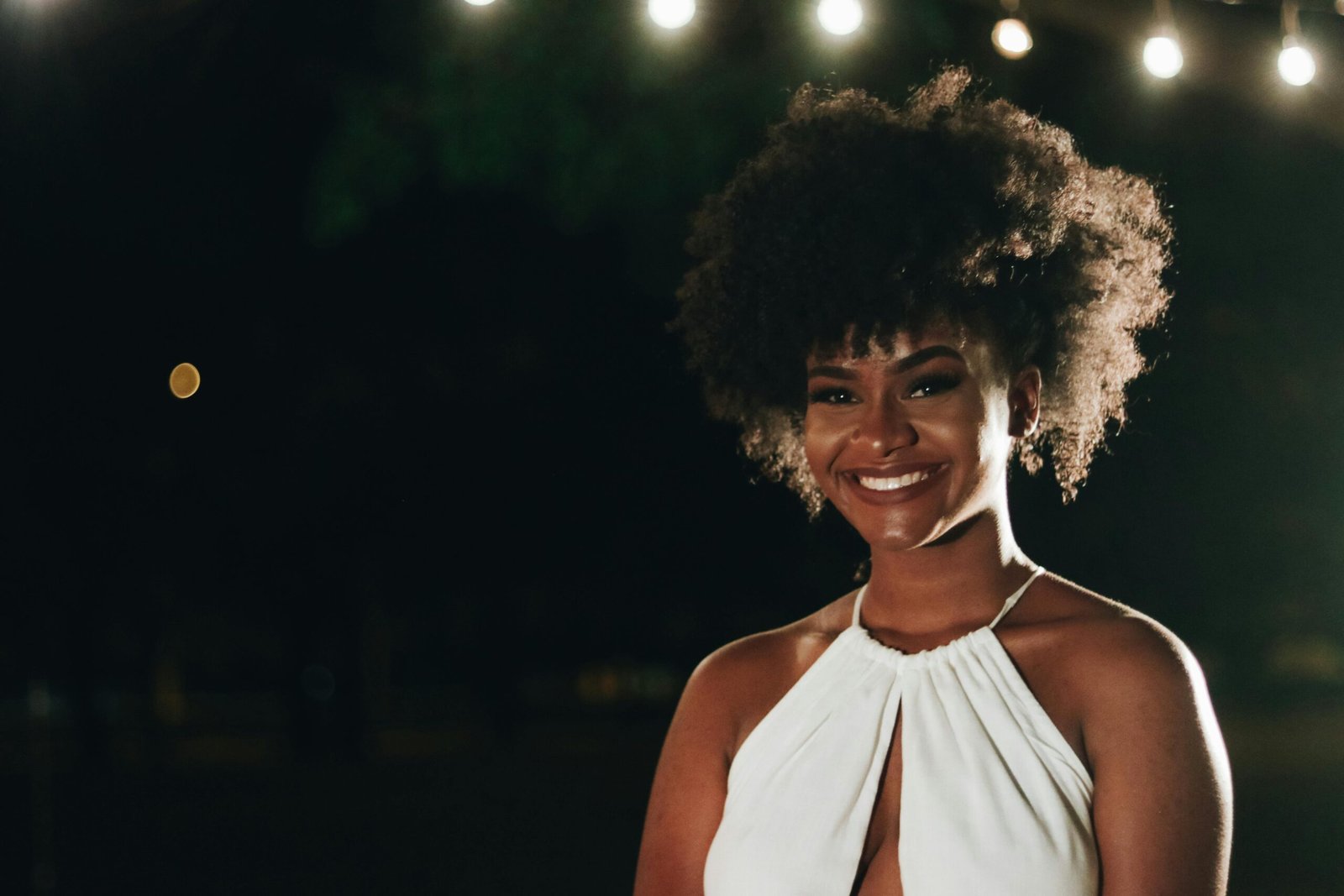 A beautiful woman with afro hair smiling outdoors at night under soft lights.